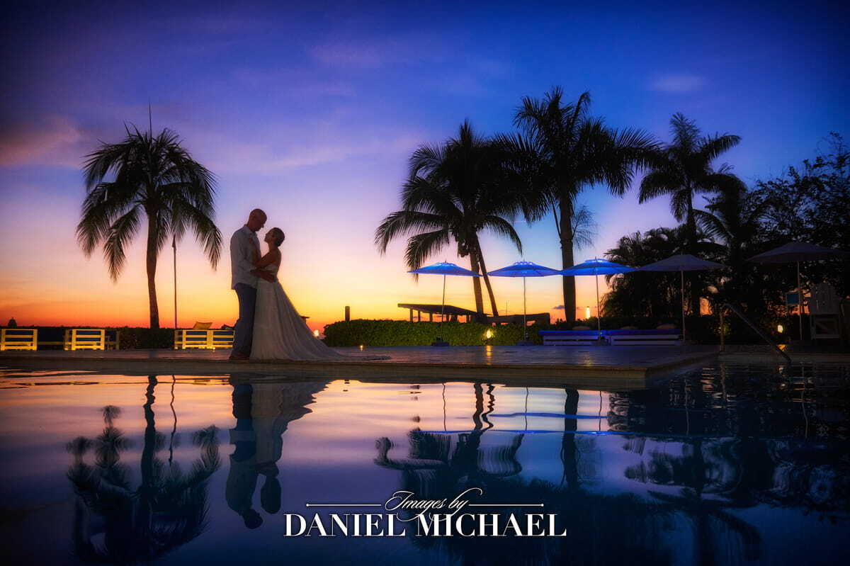 a bride and groom standing next to a pool with the sun setting in the sky behind them